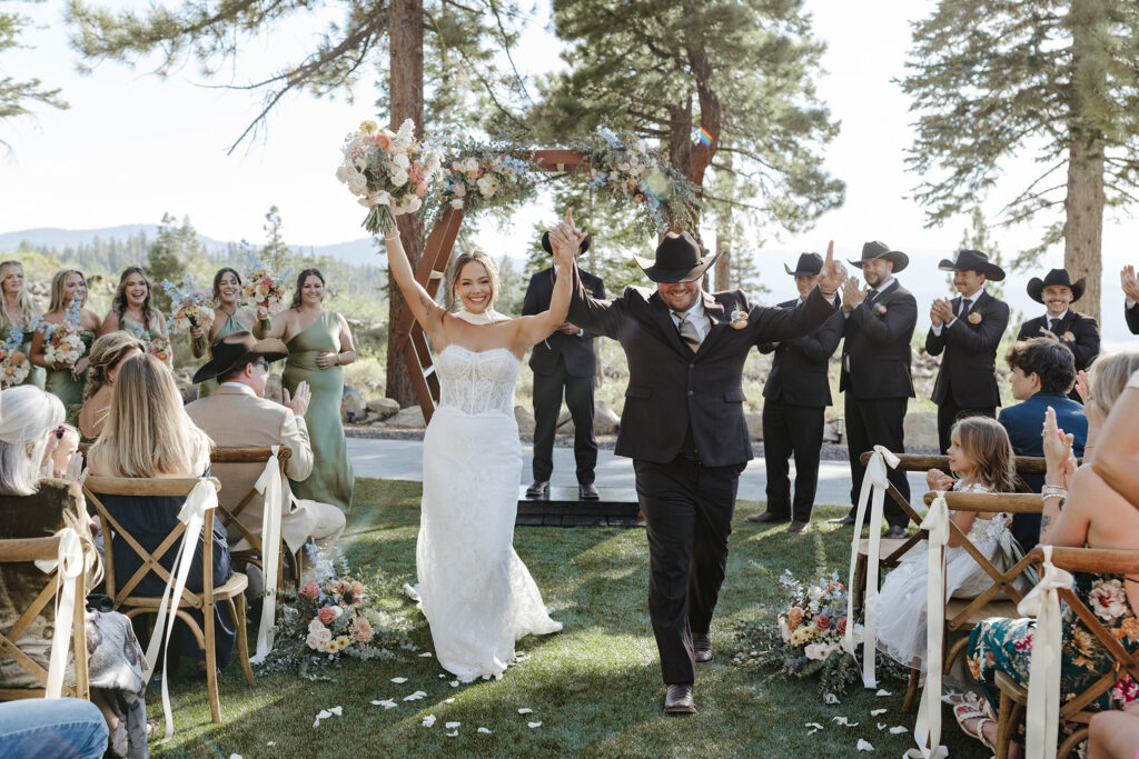 Wedding couple holding hands up in air together and smiling while walking down aisle after ceremony at Austin Ridge Truckee with guests on either side and bridal party clapping in background
