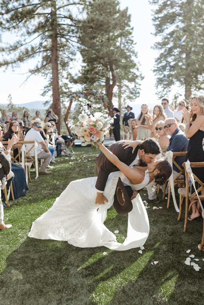 Groom leaning bride back and kissing in wedding aisle after ceremony at Austin Ridge Truckee while guests cheer in background