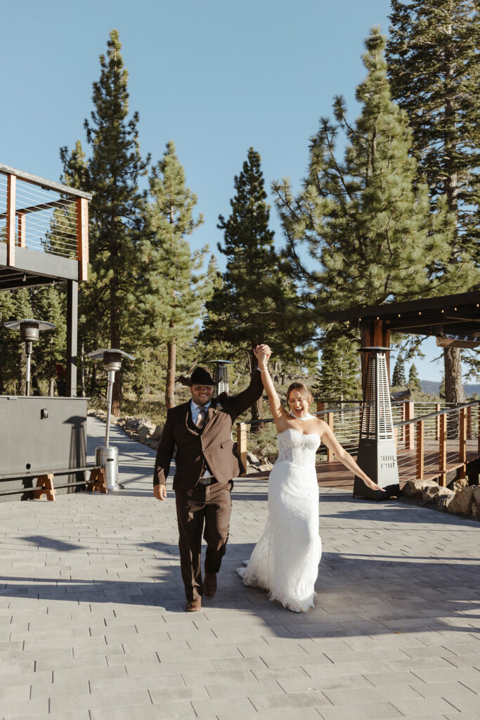 Wedding couple holding hands up in the air together and celebrating while walking into reception at Austin Ridge Truckee with pine trees in background