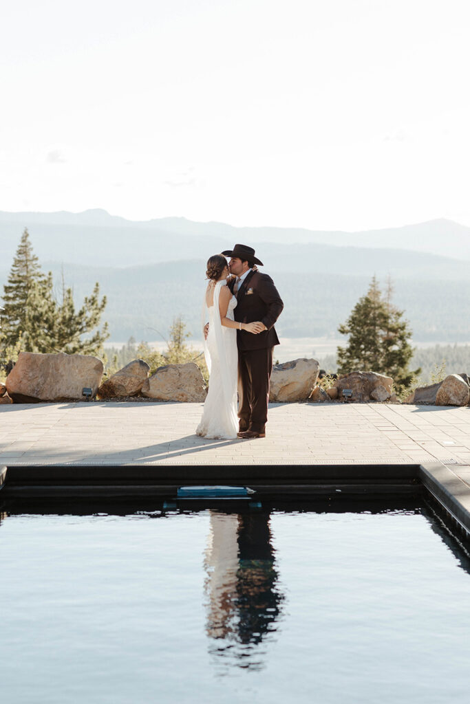 Wedding couple holding each other while groom kisses bride's cheek while standing at far end of pool with mountains in background at Austin Ridge Truckee
