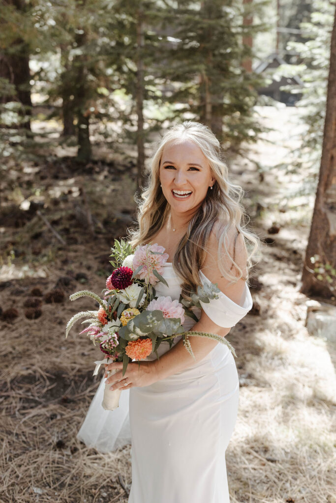 Elopement bride smiling at camera while holding floral bouquet in full wedding attire with pine trees in background at Lake Tahoe