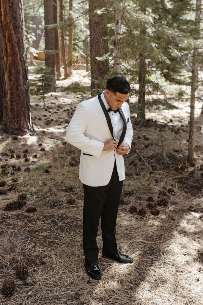 Groom looking down and buttoning wedding jacket while standing in front of pine trees at Lake Tahoe