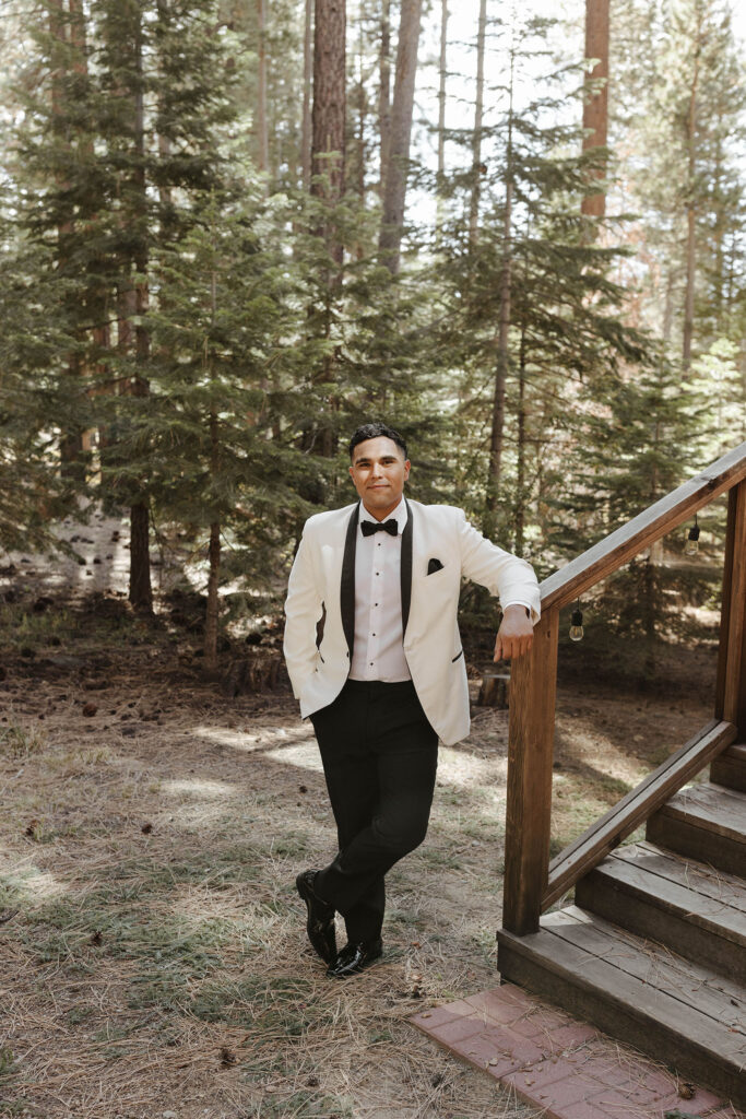 Elopement groom leaning on wooden railing while smiling at camera in full wedding attire with pine trees in background at Lake Tahoe