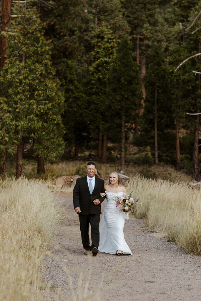 Bride holding dad's arm and smiling while walking down trail to elopement ceremony with thick pine trees in background at Lake Tahoe