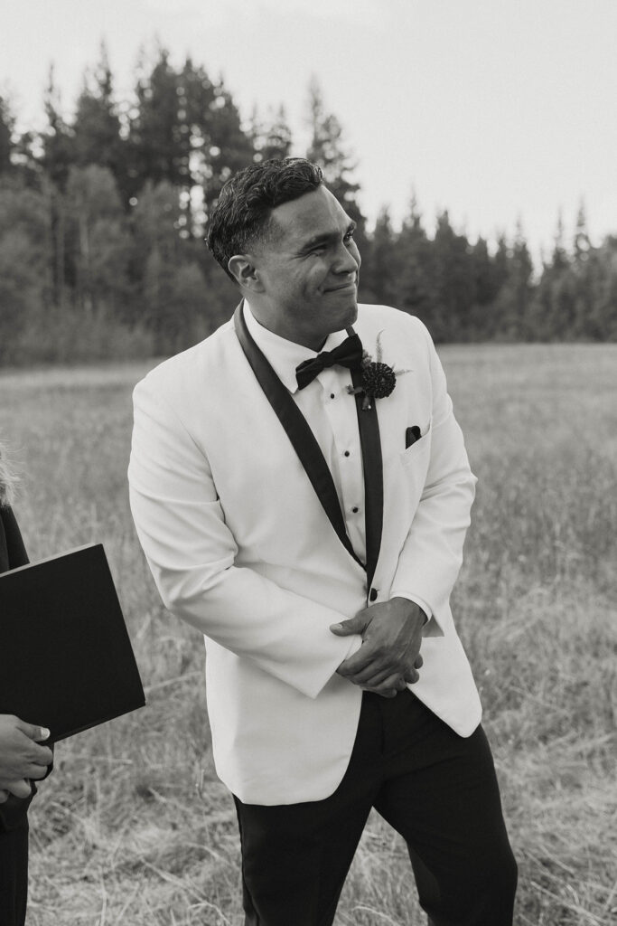 Groom smiling while standing with hands clasped and watching bride walk down to elopement ceremony with pine trees in background at Lake Tahoe
