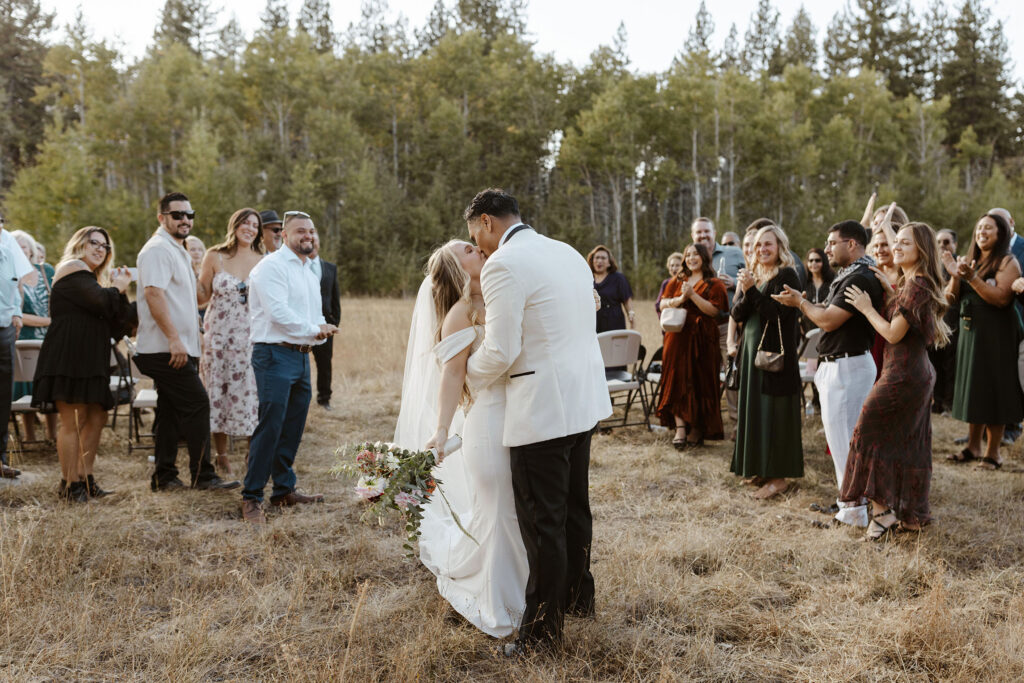 Elopement couple kissing after ceremony while bride holds colorful floral bouquet and guests clap on either side in Lake Tahoe with trees in background