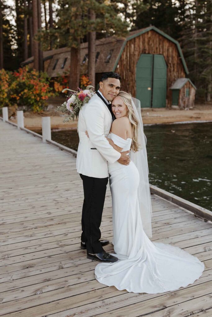 Elopement couple holding each other close while smiling at camera and standing on wooden pier together at Lake Tahoe with wooden barn and pine trees in background