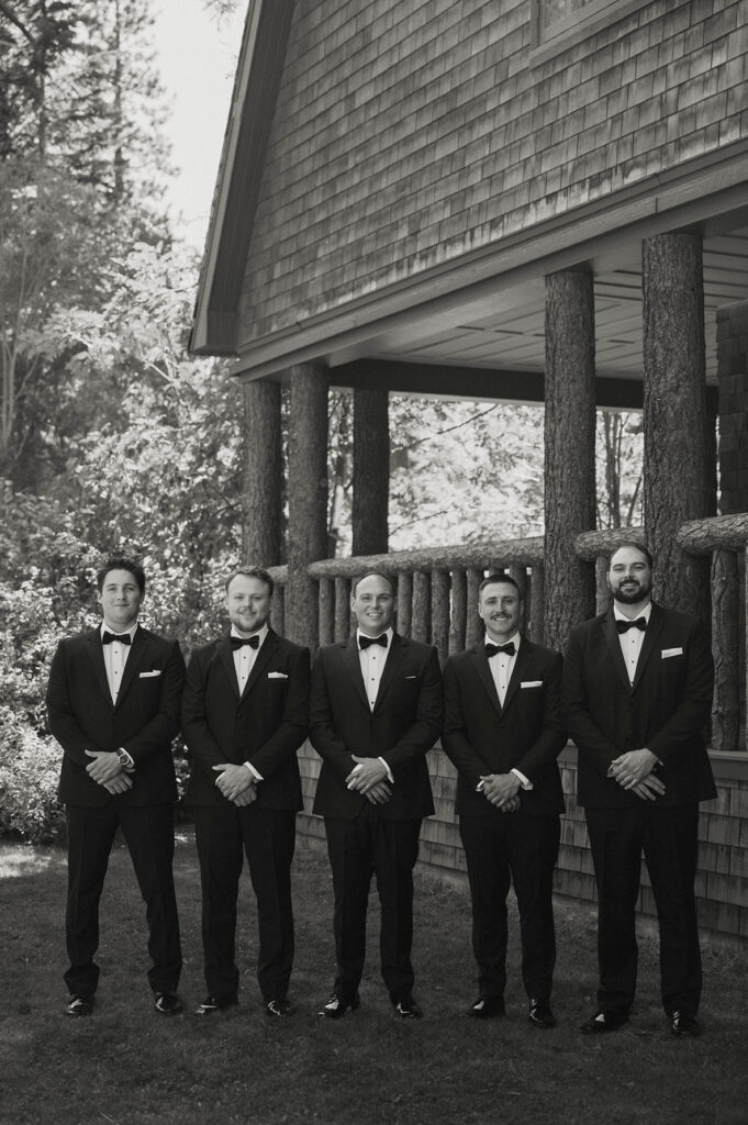 Wedding groom standing in between groomsmen in full wedding attire while standing with hands clasped and smiling at camera in front of natural wooden railing at Valhalla Tahoe