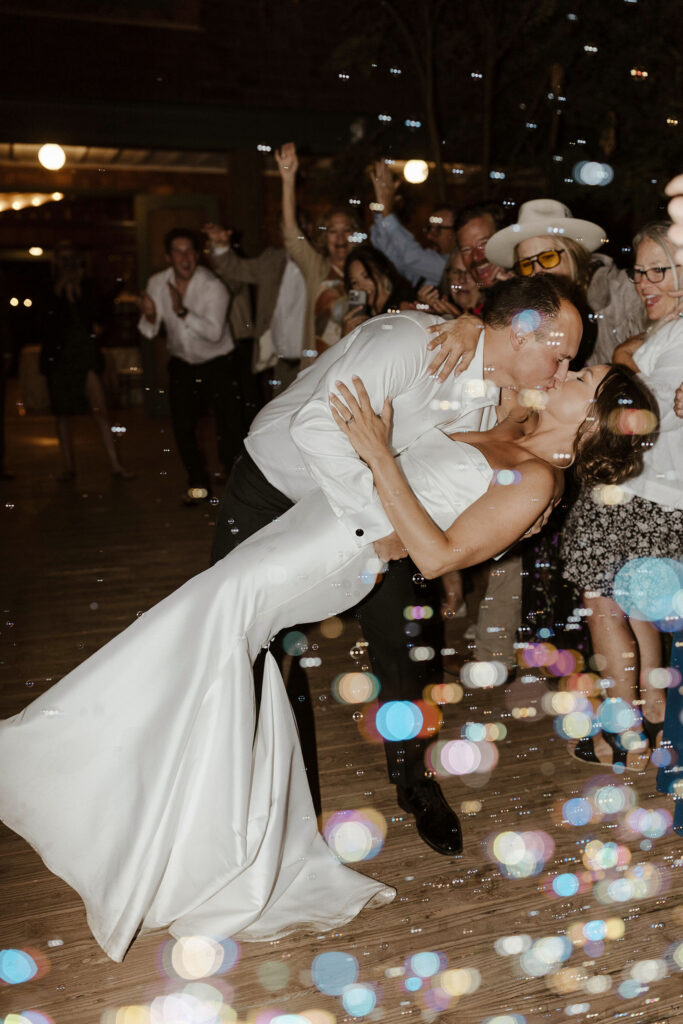 Groom leaning bride back and kissing her while wedding guests blow bubbles around them and celebrate at Valhalla Tahoe