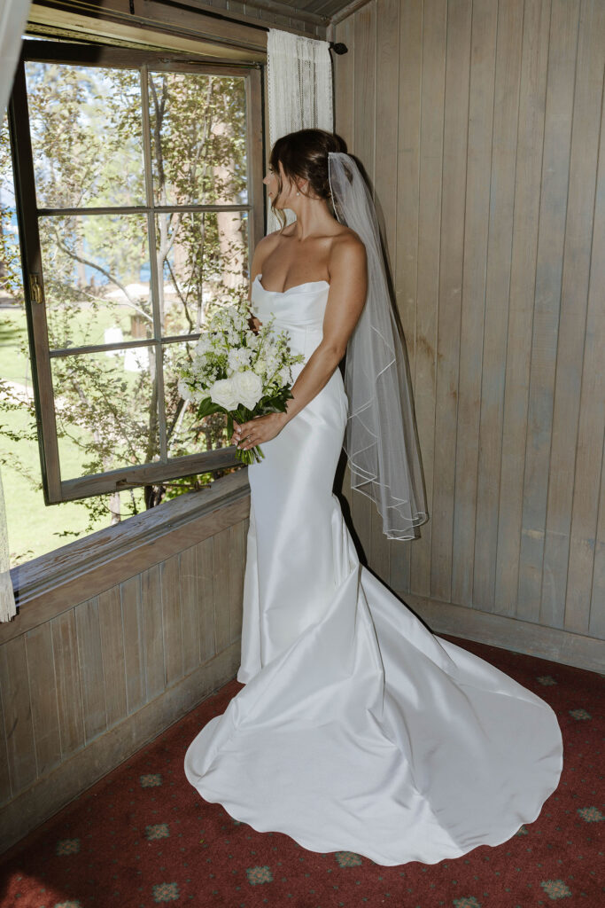 Bride in full wedding attire holding white floral bouquet while looking out large window at Valhalla Tahoe