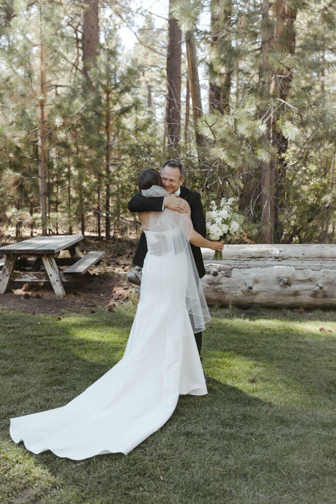 Bride's dad hugging bride while she holds floral bouquet during first look at Valhalla Tahoe with large log and pine trees in background