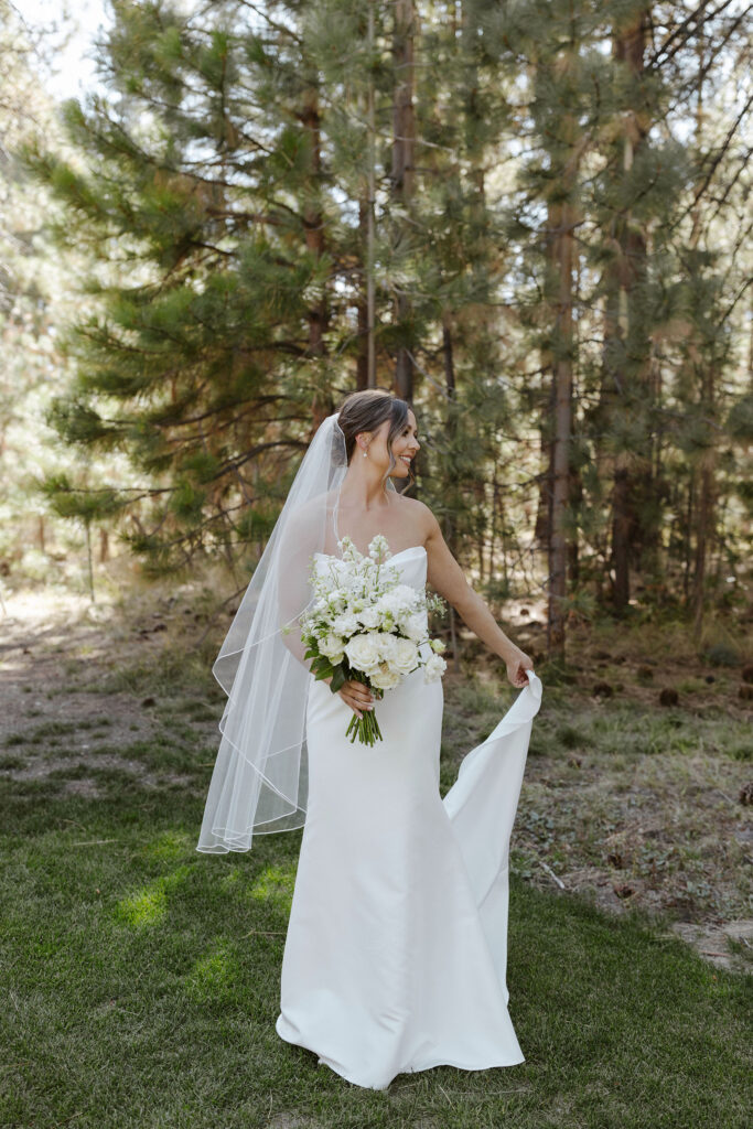 Bride holding wedding dress trail and floral bouquet while looking over shoulder and smiling at Valhalla Tahoe with pine trees in background