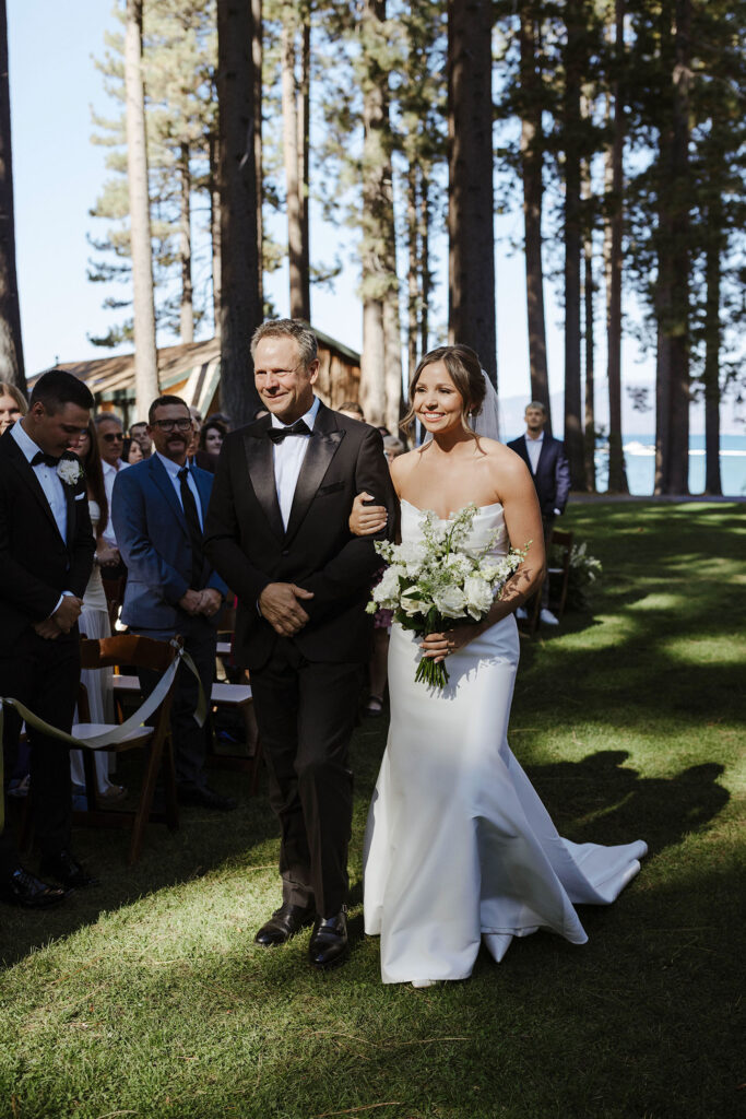 Bride holding dad's arm and smiling while walking down wedding aisle with guests on either side to ceremony at Valhalla Tahoe