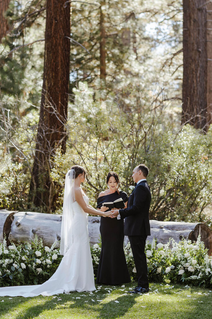 Wedding couple holding hands while officiant reads vows during ceremony in front of white flowers and greenery at Valhalla Tahoe