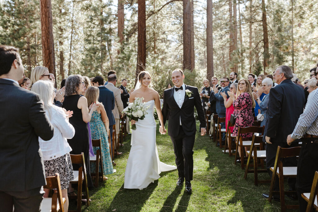 Wedding couple holding hands and walking down aisle together after ceremony while smiling at camera with guests clapping on either side at Valhalla Tahoe
