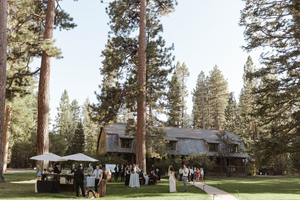 Overview of cocktail party after wedding ceremony on grass in front of tall trees and wooden building at Valhalla Tahoe