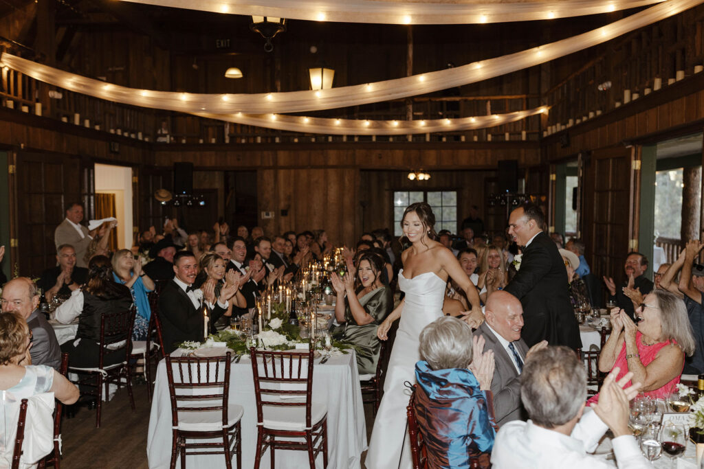 Wedding couple holding hands and smiling while walking through reception full of guests cheering with string lights hanging above them at Valhalla Tahoe