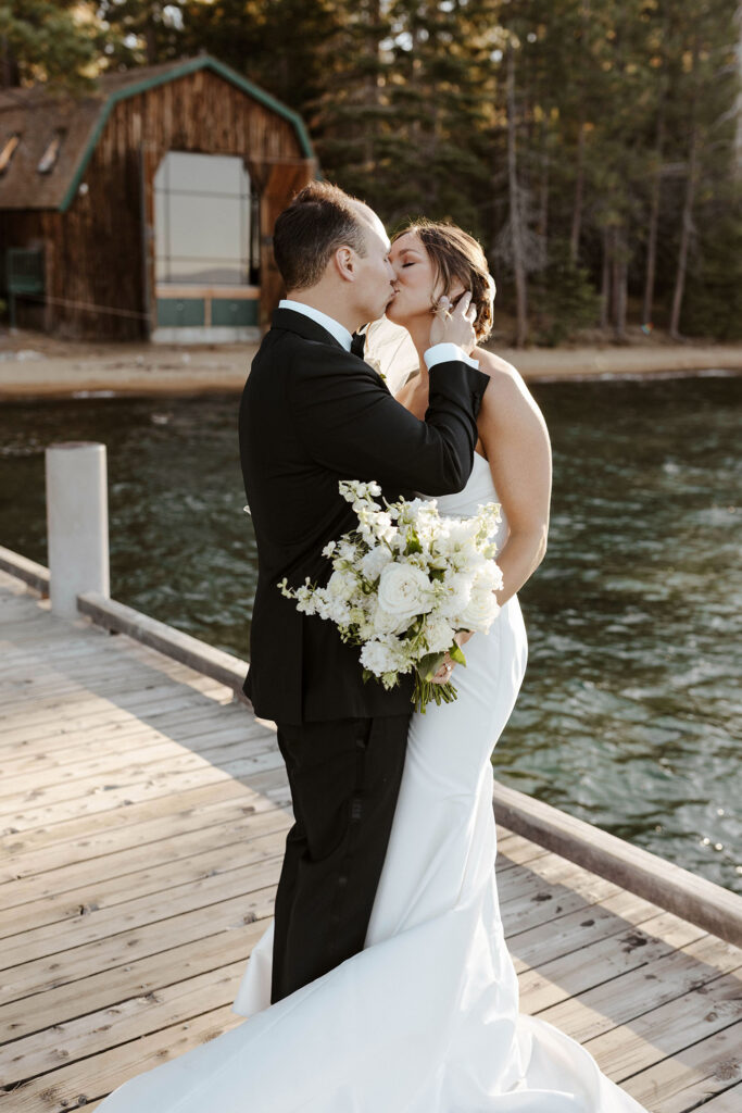 Wedding couple kissing while on a pier together with water and wooden building in background at Valhalla Tahoe
