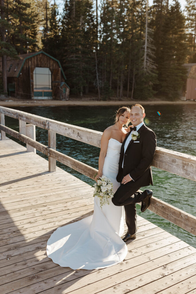 Wedding couple leaning on wooden railing together on a pier and smiling at camera with Lake Tahoe and pine trees in background