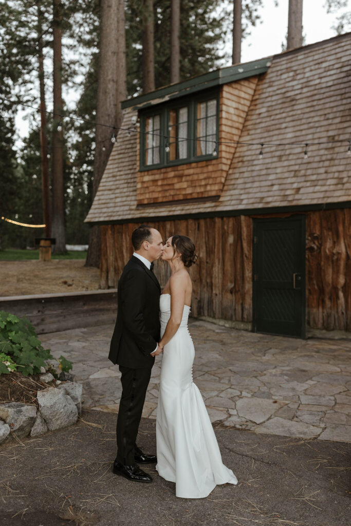 Wedding couple holding hands and kissing in front of wooden building and tall pine trees at Valhalla Tahoe