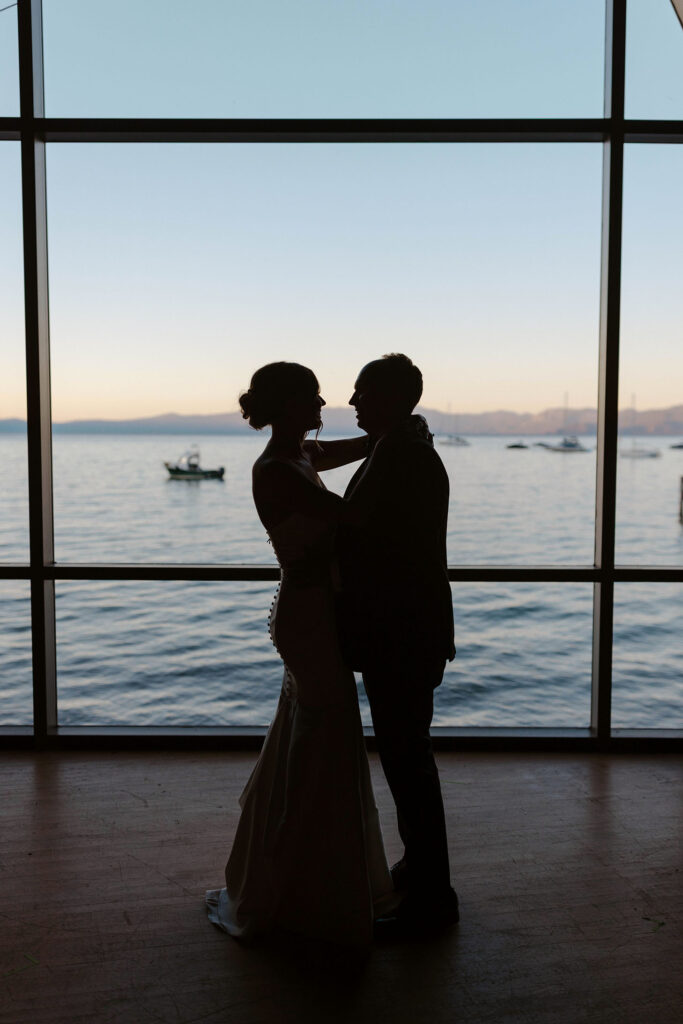 Silhouettes of wedding couple standing together in front of large windows with Lake Tahoe in background at Valhalla