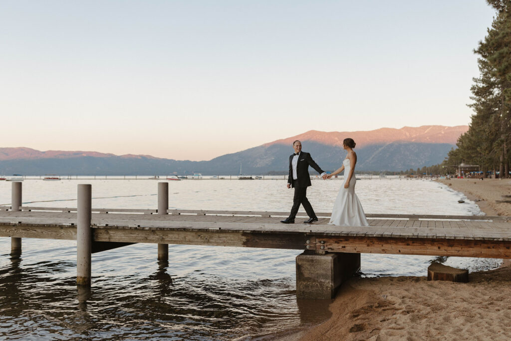 Groom holding bride's hand and leading her along wooden pier on Lake Tahoe with mountains in background