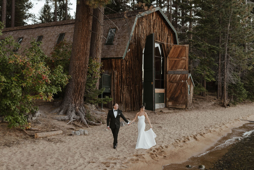 Wedding couple holding hands while walking along sandy beach together and looking at Lake Tahoe with wooden barn and lots of trees in background