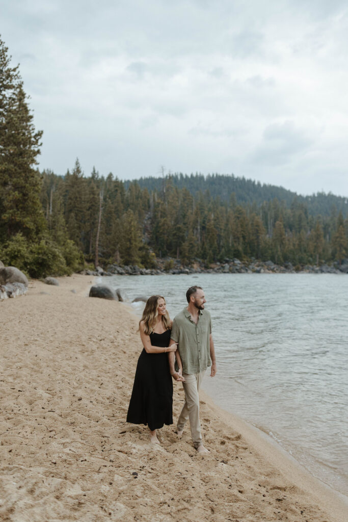 couple walking on the beach taking engagement photos in the rain in lake tahoe, looking into the distance