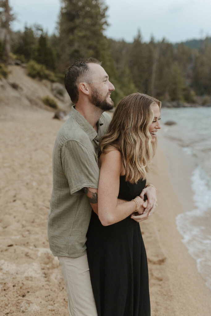 man hugging woman from behind while she laughs on a beach in lake tahoe
