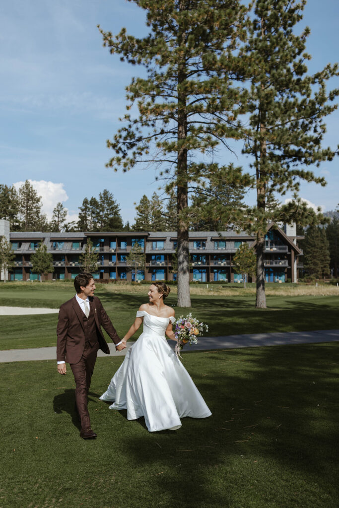 Wedding couple holding hands while walking across grass field and smiling at each other in front of building at Edgewood Tahoe