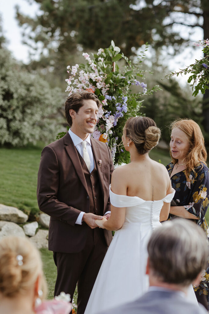 Wedding couple holding hands and smiling at each other during ceremony at Edgewood Tahoe with colorful florals on arch behind them