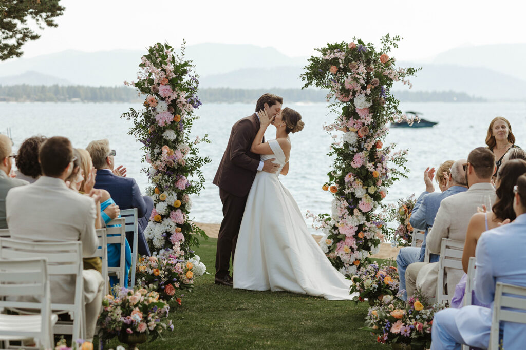 Wedding couple's first kiss in between pink and white floral columns during ceremony at Edgewood while guests clap with Lake Tahoe in background