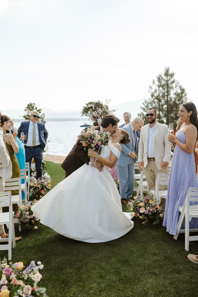 Groom leaning bride back and kissing her in wedding aisle with guests on either side and Lake Tahoe in background at Edgewood