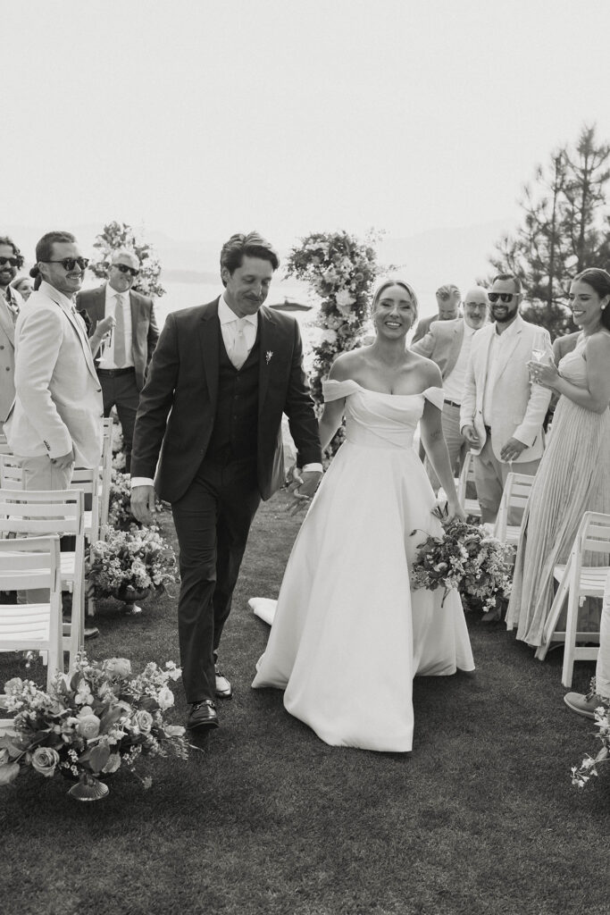 Wedding couple holding hands and walking down aisle together after ceremony with guests on either side smiling at them and Lake Tahoe in background at Edgewood