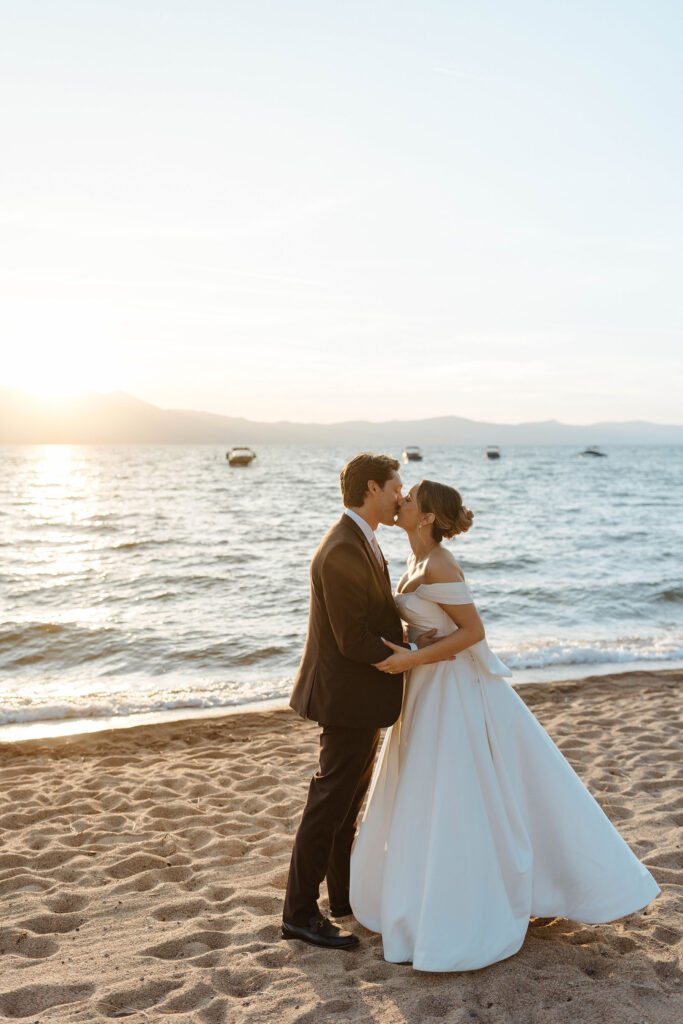 Wedding couple kissing and holding each other while on a beach at sunset with Lake Tahoe in background at Edgewood