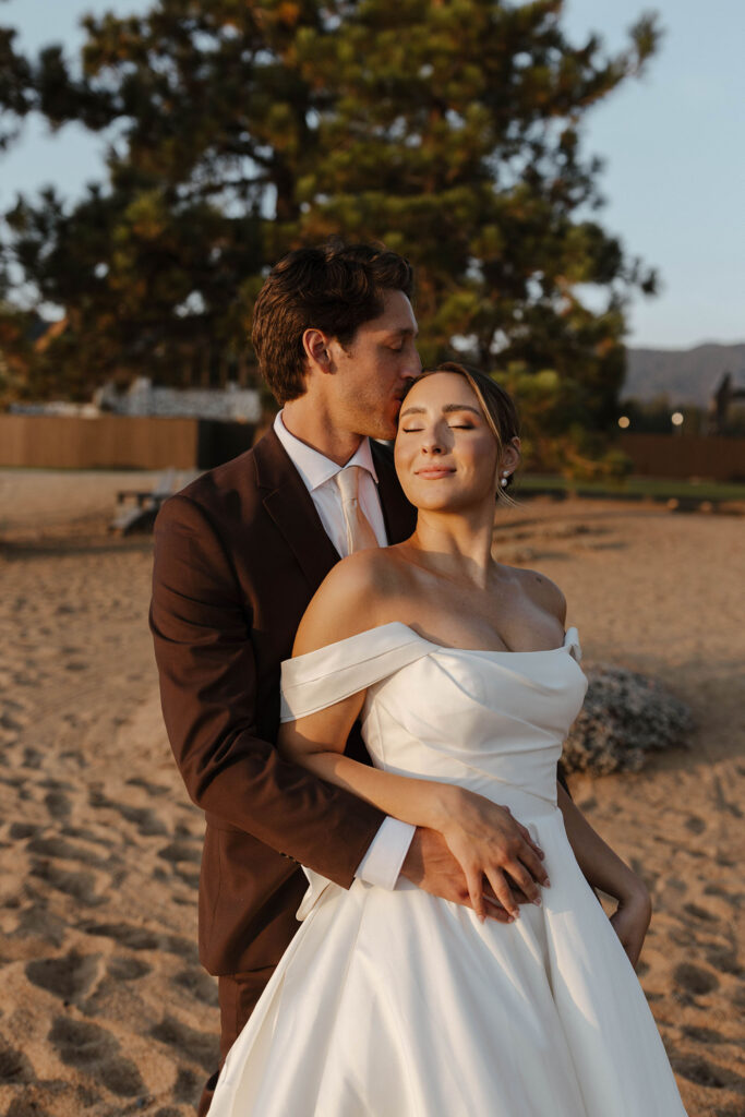 Groom hugging wedding bride from behind while holding hands on a beach at Edgewood Tahoe with trees in background