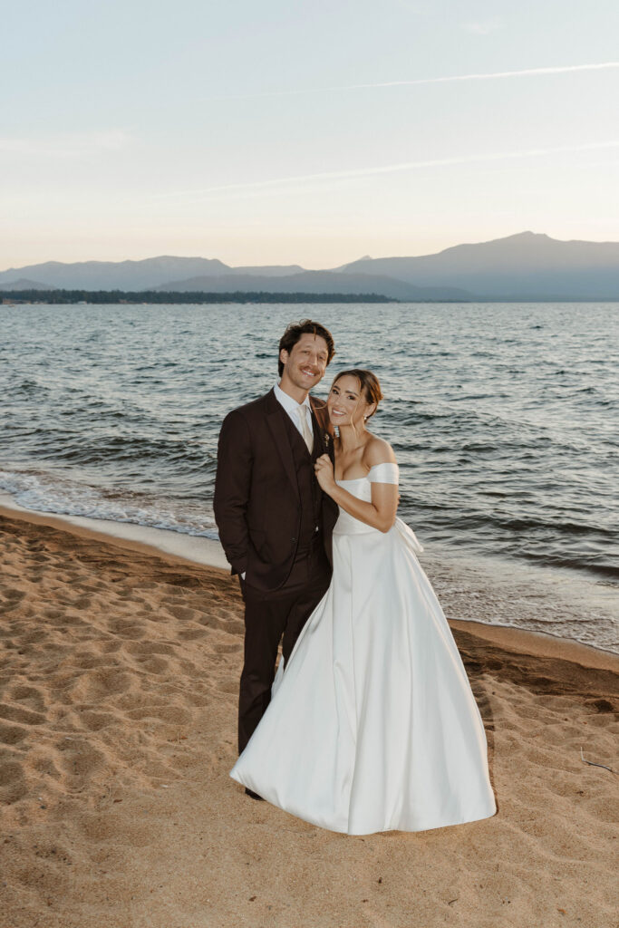 Wedding couple holding each other while smiling at camera and standing on beach together at Edgewood with Lake Tahoe in background