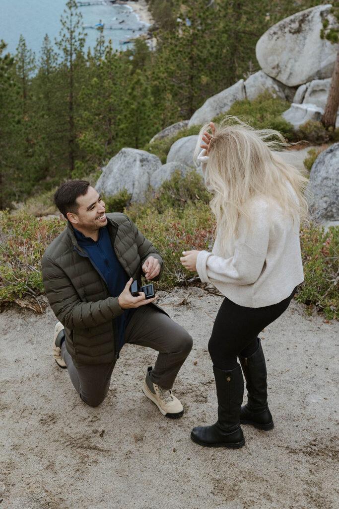 Man on one knee smiling and proposing holding engagement ring box with large rocks and trees in background in Lake Tahoe