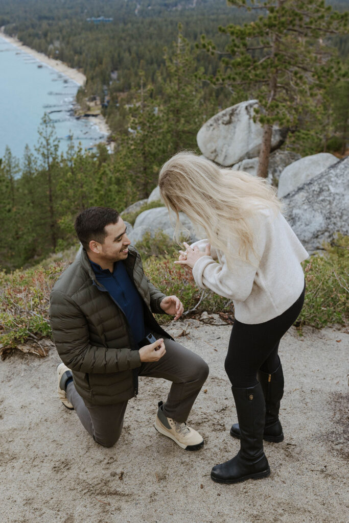 Man on one knee while woman slides engagement ring onto finger with large rocks and trees in background in Lake Tahoe
