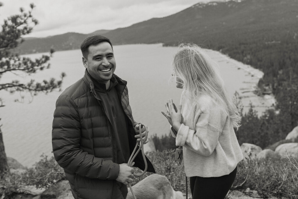 Man smiling at camera while standing next to fiancé with Lake Tahoe and forest of trees in background