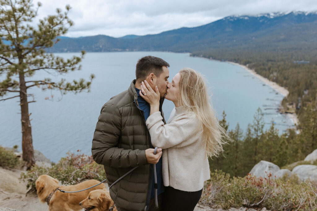 Newly engaged couple kissing while woman holds fiancé's face with engagement ring on finger and Lake Tahoe and mountains in background