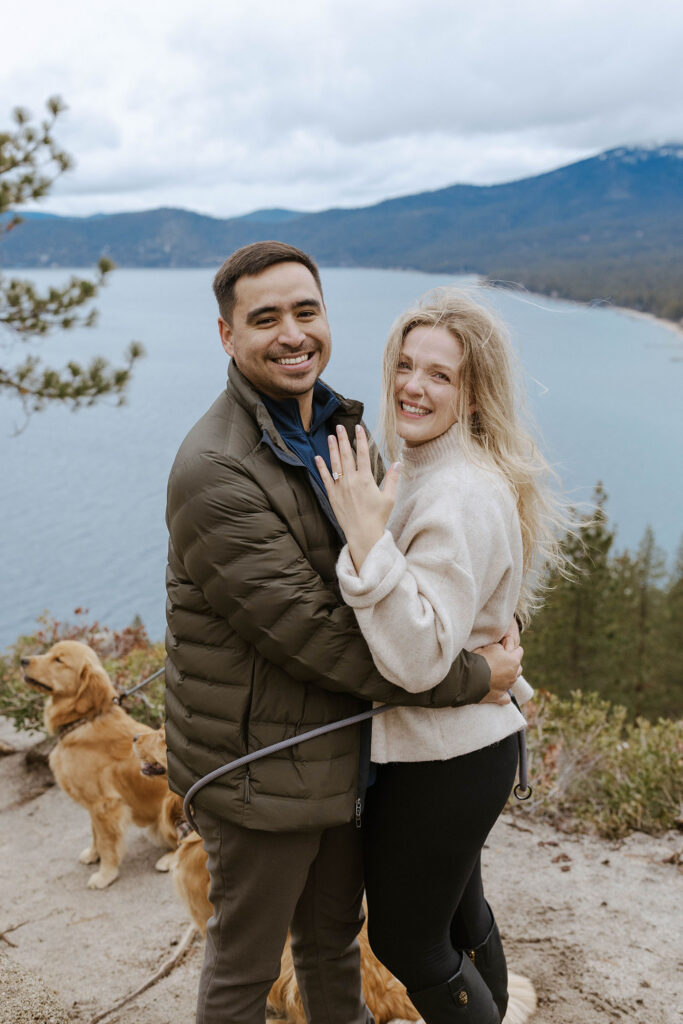 Woman showing off engagement ring on hand while couple hugs each other and smiles at camera with Lake Tahoe in background