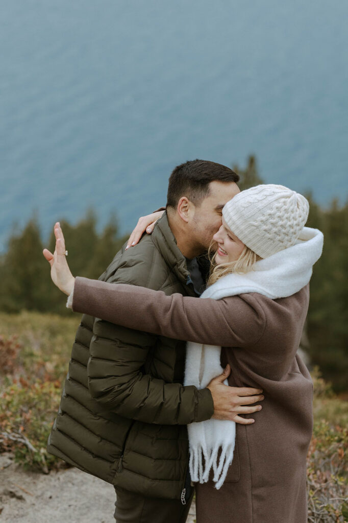 Woman holding hand out and looking at engagement ring on finger while fiancé hugs her with Lake Tahoe in background
