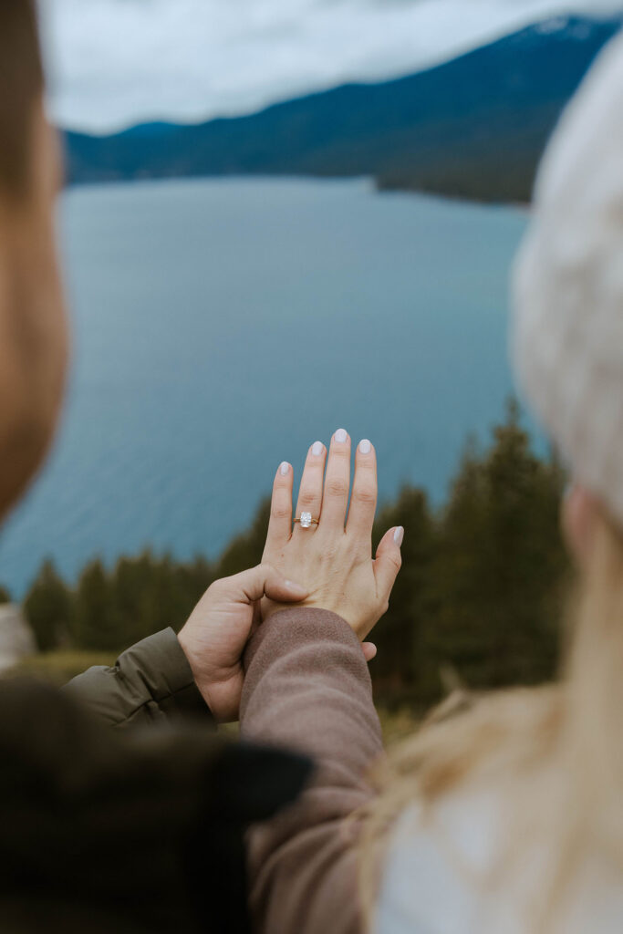 Close up of woman's hand with engagement ring and fiancé holding her hand with Lake Tahoe in background
