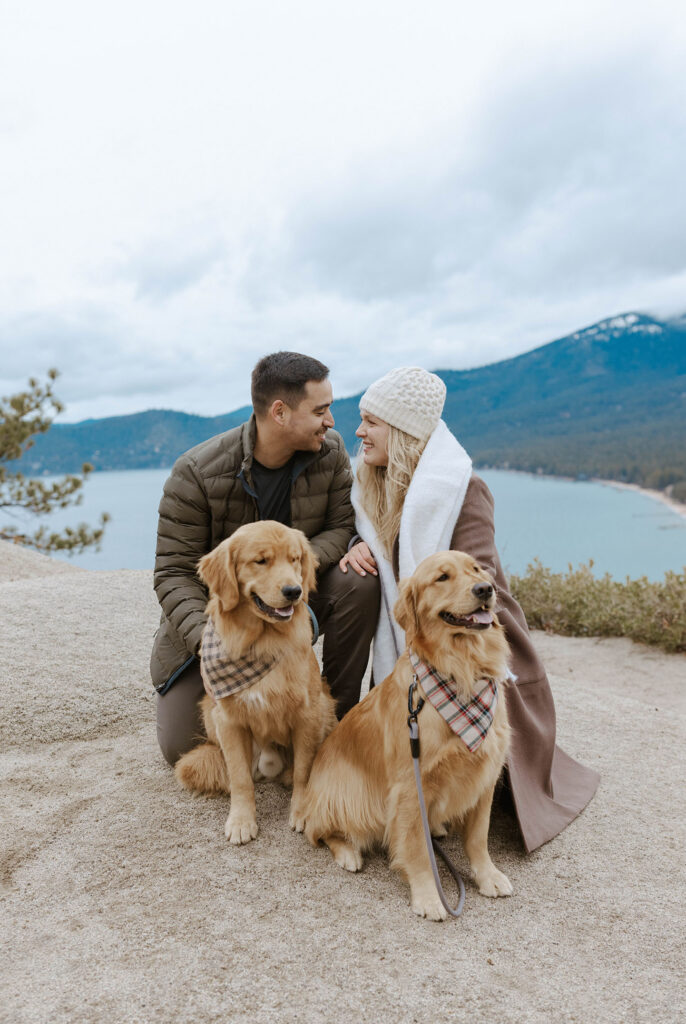 Engagement couple crouching together next to their two golden retrievers and smiling at each other with Lake Tahoe and mountains in background