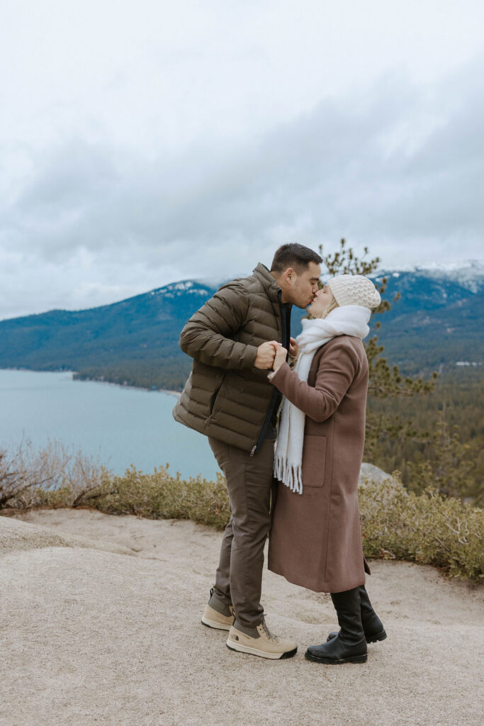 Engagement couple holding hands and kissing on trail with Lake Tahoe and mountains in background