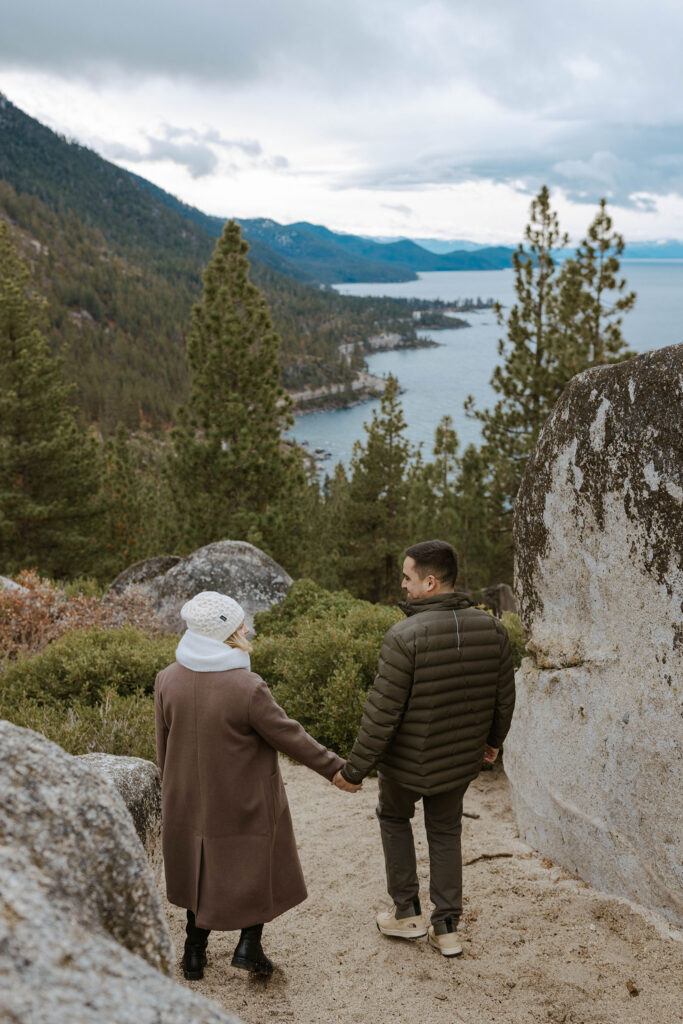 Engagement couple holding hands while walking down trail together next to large rocks towards tall trees and Lake Tahoe in distance