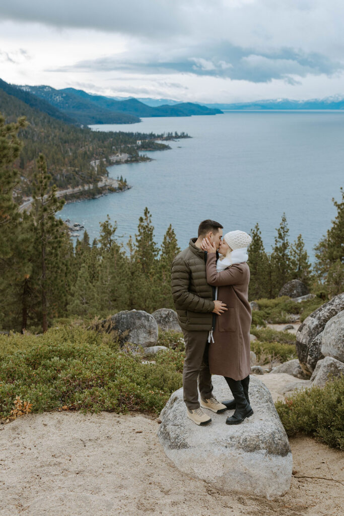 Engagement couple holding each other and kissing while standing on rock together with tall trees and Lake Tahoe in background
