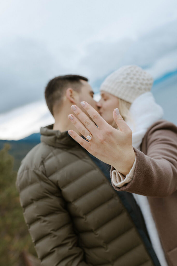 Engagement couple kissing while woman holds hand with engagement ring close to camera in Lake Tahoe