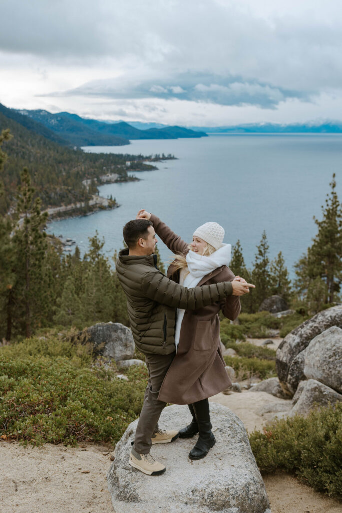 Engagement couple holding hands while dancing together on a rock with tall trees and Lake Tahoe in background